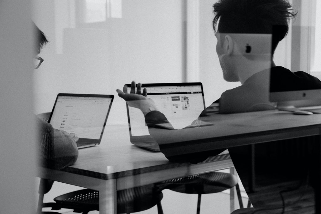A black and white photo of two people in an office looking at their computers having a discussion