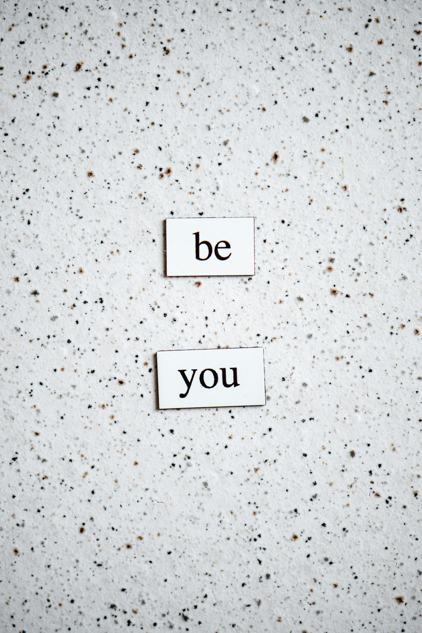 A sprinkled pattern white background and two words written in black, Be You.
