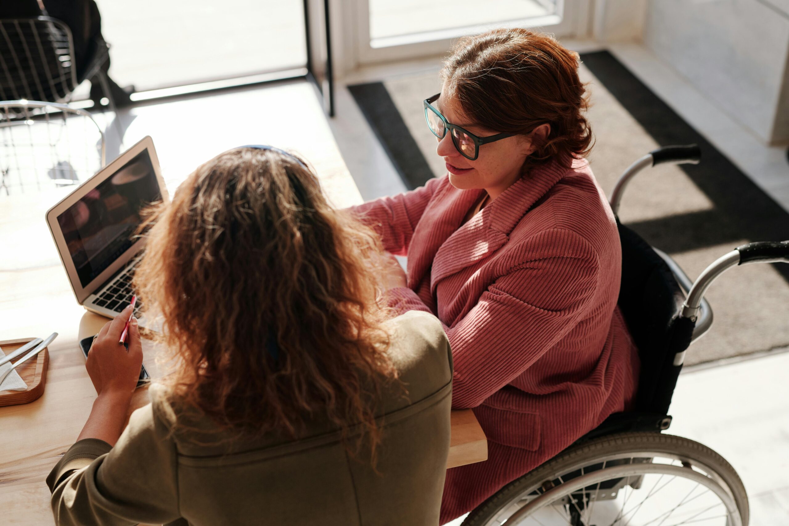 Two women sit side by side at a coffee shop as the sunlight shines on the table giving a warm feeling.